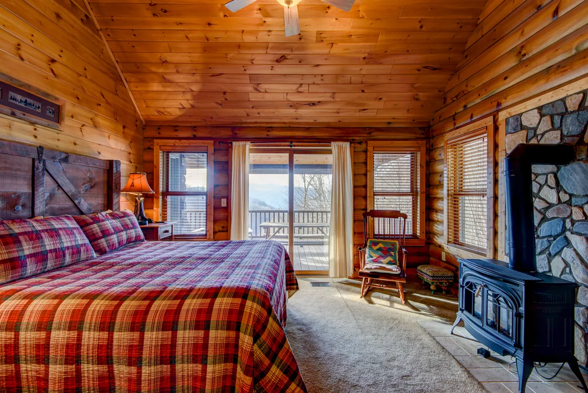 A plush king bed with a plaid bedspread anchors the room, complemented by a stone fireplace, large windows revealing woodland views, and a wooden ceiling overhead. The atmosphere is warm and tranquil, suggesting a private retreat ideal for unwinding.