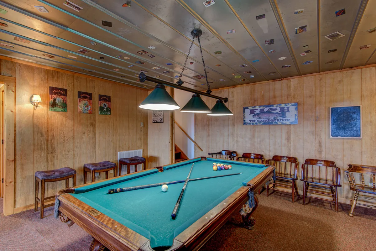 The scene shows a vibrant game area with a pool table under hanging lights, surrounded by wooden chairs and decorated with playful posters. The setting invites guests to enjoy friendly competition and celebrate together, reflecting a casual, youthful vibe.