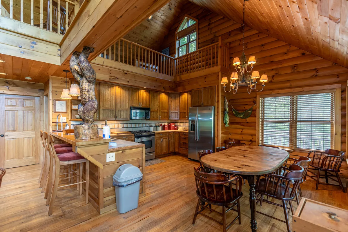 The image showcases a central dining table near a kitchen island, with warm wooden textures and large windows providing abundant light and views of nature. The setup encourages group meals and easy flow between dining and kitchen spaces, with a welcoming, communal feel.