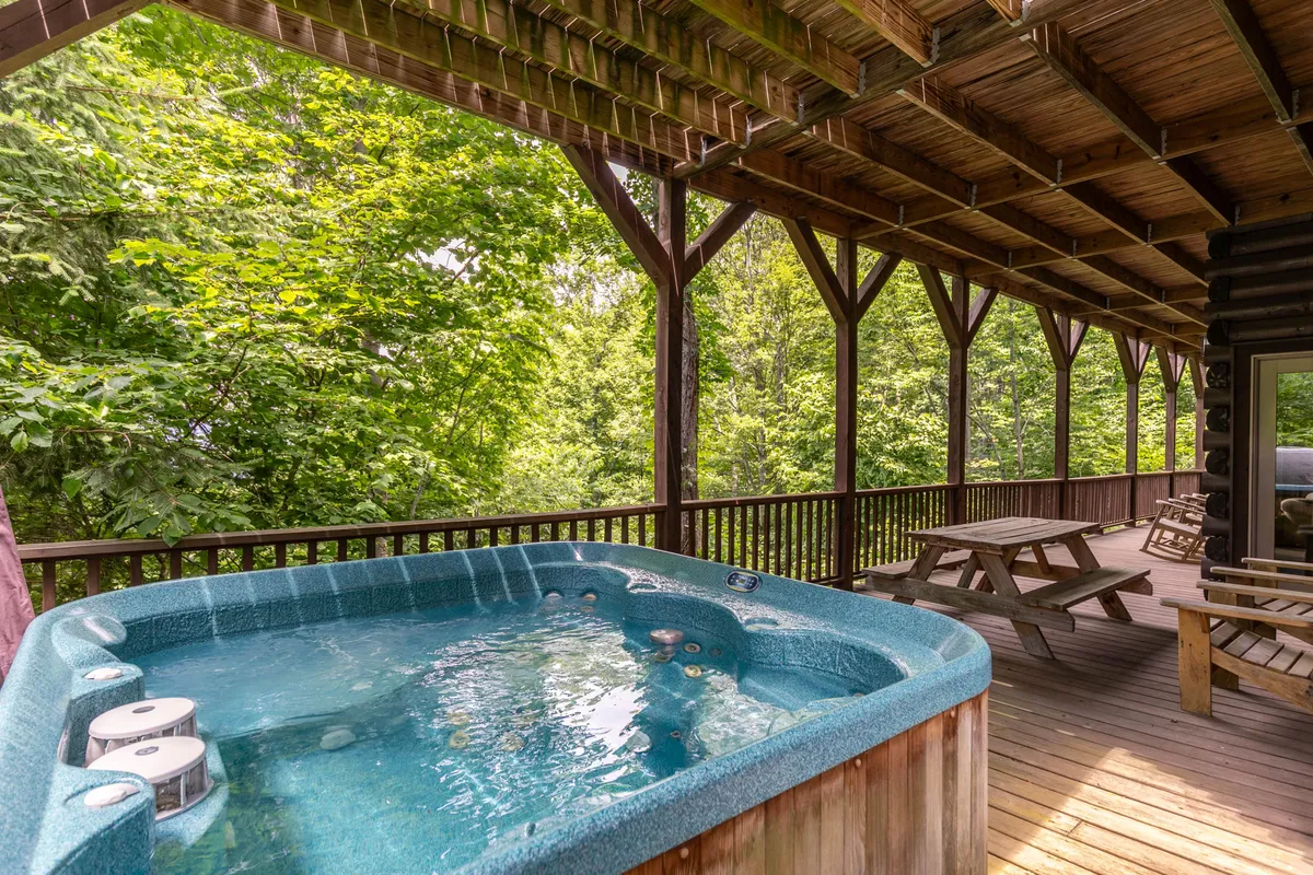 This image captures the lower deck of the property, highlighting a bubbling hot tub surrounded by lush greenery. A picnic table and wooden deck space are visible, inviting group gatherings and outdoor dining. The tranquil setting suggests a place for relaxation and connection with nature.