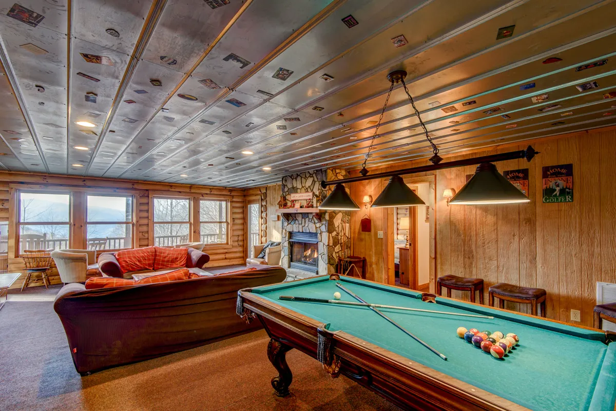 This image captures a spacious game room interior featuring a pool table as the focal point, with a cozy sofa positioned near a gas log fireplace. Large windows flood the room with natural light, and the seating area encourages socializing and relaxation. The ambiance suggests both active game nights and tranquil lounging.
