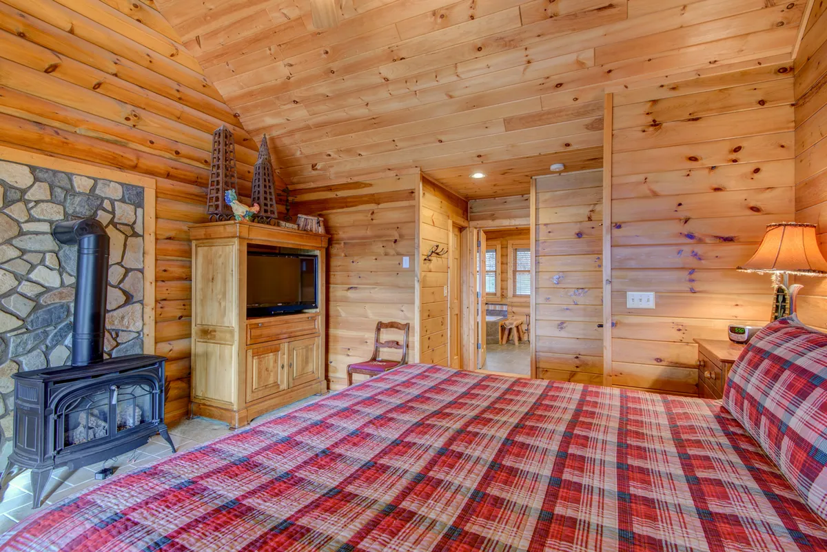 A cozy adjacent bedroom with plaid bedding, a stone fireplace, and a television unit. Wooden walls and comfortable furnishings create a warm, inviting atmosphere suitable for unwinding after a night of entertainment.