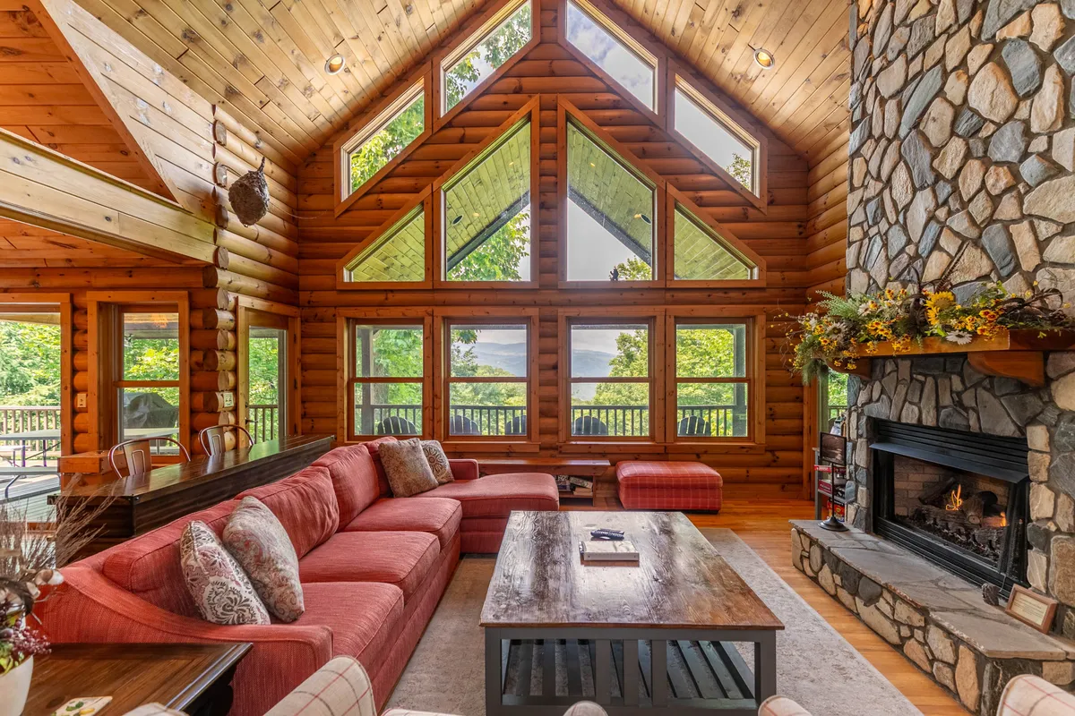 This image features a spacious great room with a large stone fireplace as the focal point, surrounded by an inviting red sectional sofa. Expansive windows fill the room with natural light and frame stunning mountain views, while the wooden ceiling and log walls add warmth and rustic charm.