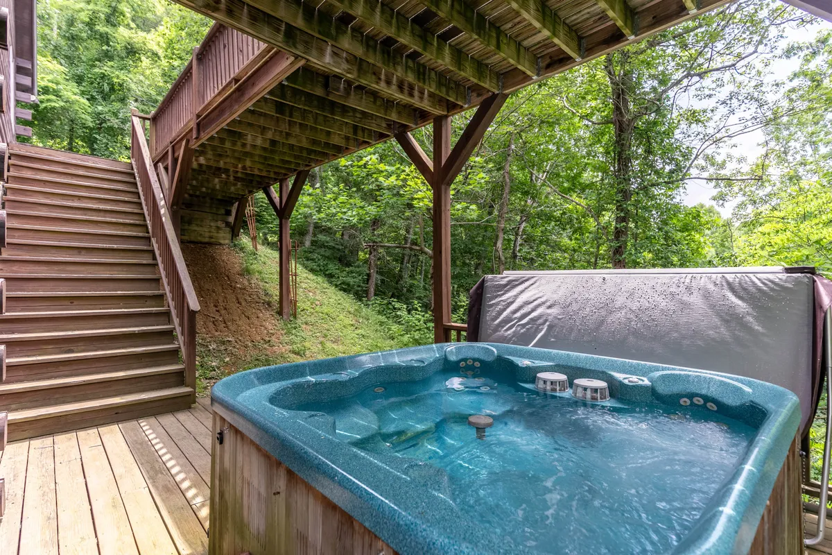 This image shows a serene hot tub on an outdoor deck surrounded by lush greenery, with wooden stairs leading up and forested views in the background. The setup is inviting for groups to unwind outside, emphasizing a natural, private setting.