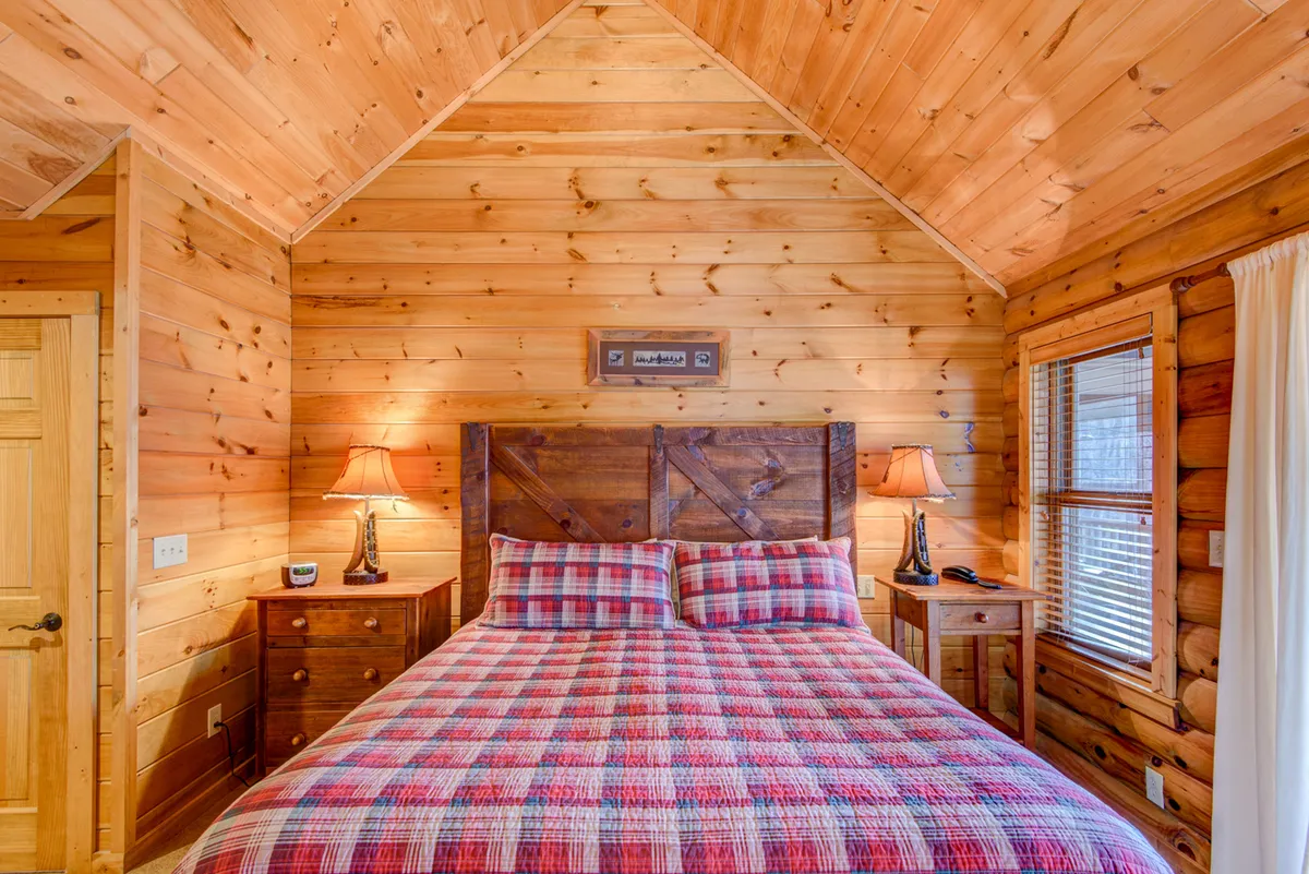 A cozy bedroom featuring a plaid bedspread, warm wooden walls, a nightstand with a glowing lamp, and simple framed artwork. The room exudes comfort and warmth, suitable for restful nights.
