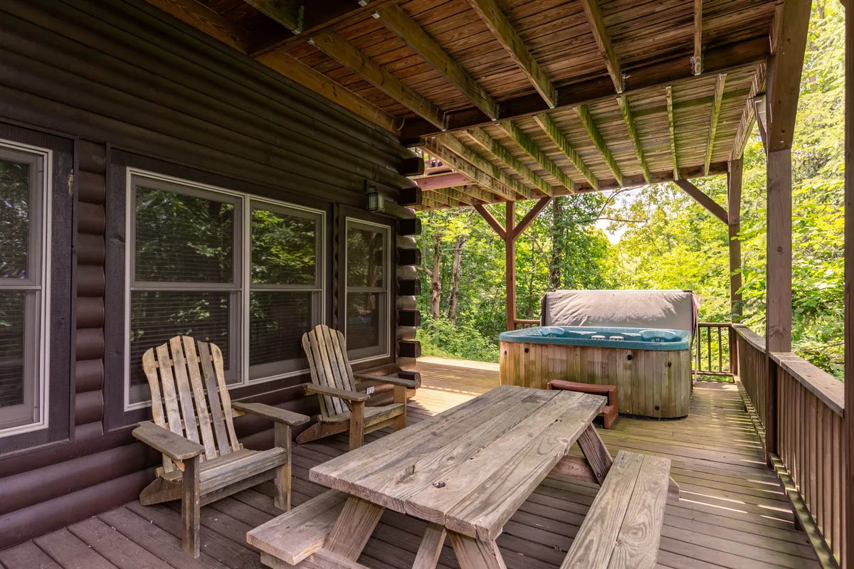 A wooden deck featuring comfortable outdoor seating, a dining table, and a hot tub, all set amid lush forest greenery. The deck offers panoramic views, suggesting opportunities for relaxation, stargazing, and soaking in the beauty of the mountains after a day of exploration.