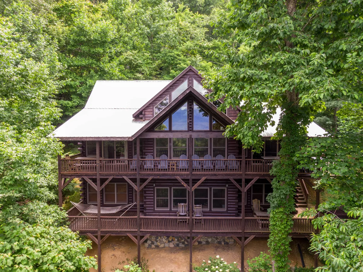 The image showcases a charming log cabin nestled among tall trees, with expansive windows and a spacious deck furnished with classic rocking chairs. The setting bathes in warm light, hinting at sunset views over the surrounding Blue Ridge Mountains. The deck invites relaxation, suggesting moments of sipping morning coffee or gathering with loved ones to enjoy the tranquil mountain air.