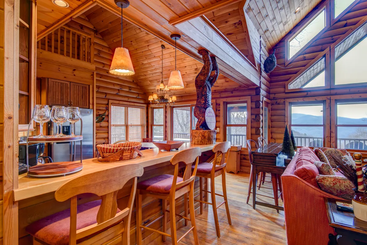 A rustic interior scene featuring a wooden bar with stools, large windows revealing mountain views, a wooden bear sculpture, and cozy lighting. The bar area is designed for social gatherings and casual entertainment, encouraging laughter and camaraderie.