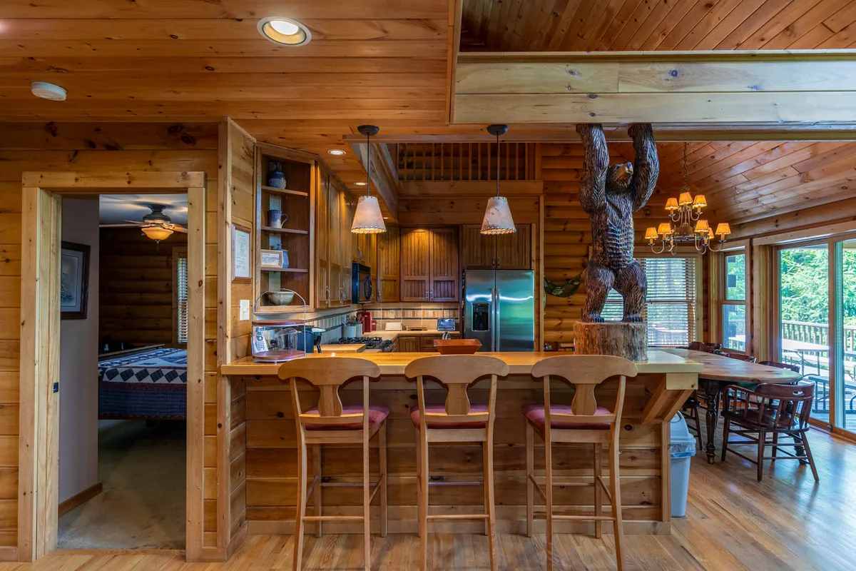 A cozy kitchen scene with a wooden bar counter and stools, kitchen appliances, a distinctive wooden bear carving, and large windows that overlook mountain scenery. The space feels rustic yet inviting, perfect for group cooking sessions and socializing.