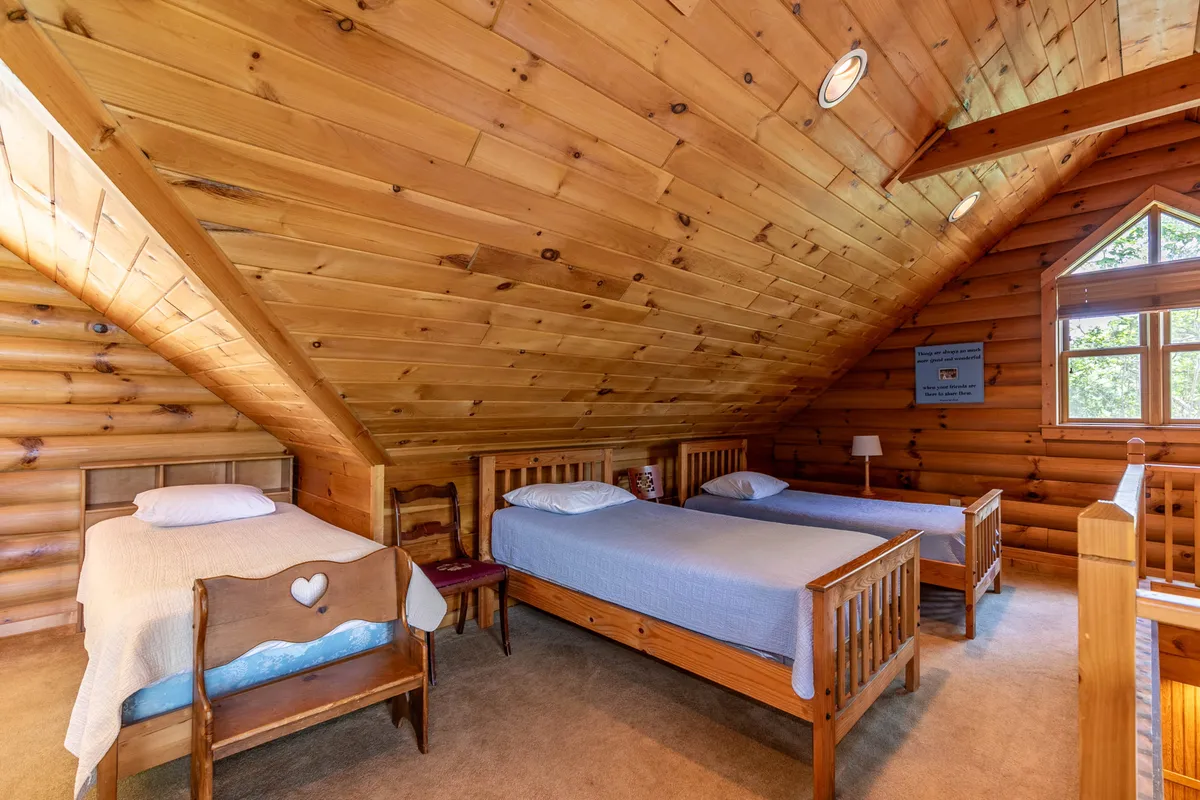A bright, lofted space with exposed wooden beams, a dramatic triangular window framing the outdoors, and multiple twin beds arranged for fun sleepovers. The room is flooded with morning light, making it feel airy and inviting, and is ideal for kids or groups of friends.
